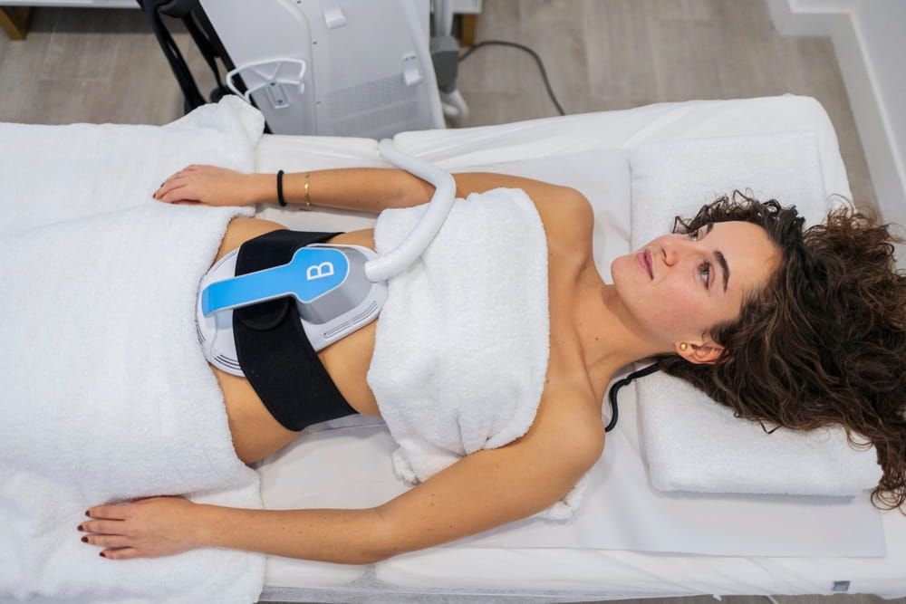 Woman lying on a bed receiving an aesthetic medicine treatment on her abdomen, targeting fat reduction and body contouring with modern technology in a clinic